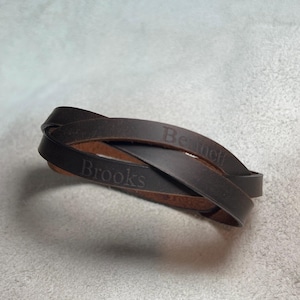 May include: A dark brown leather wrap bracelet featuring two overlapping bands. The leather has a smooth, matte finish. The name Brooks is embossed on one band, and the name Bennett is embossed on the other band in a simple, serif font.