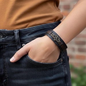 Puede incluir: Una pulsera de cuero marrón oscuro con un diseño en forma de corazón de color bronce. La pulsera se lleva en la muñeca, con la persona que lleva vaqueros azules y una camisa marrón. La pulsera mide aproximadamente 2,5 cm de ancho.