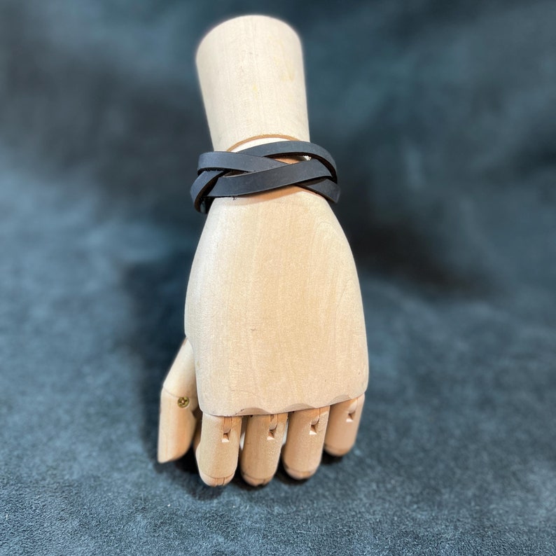 May include: A matte black leather wrap bracelet featuring multiple thin, flat strands that overlap to create a layered appearance. The bracelet is secured around a light-toned wooden mannequin hand, showcasing a simple, clean design with no visible hardware.