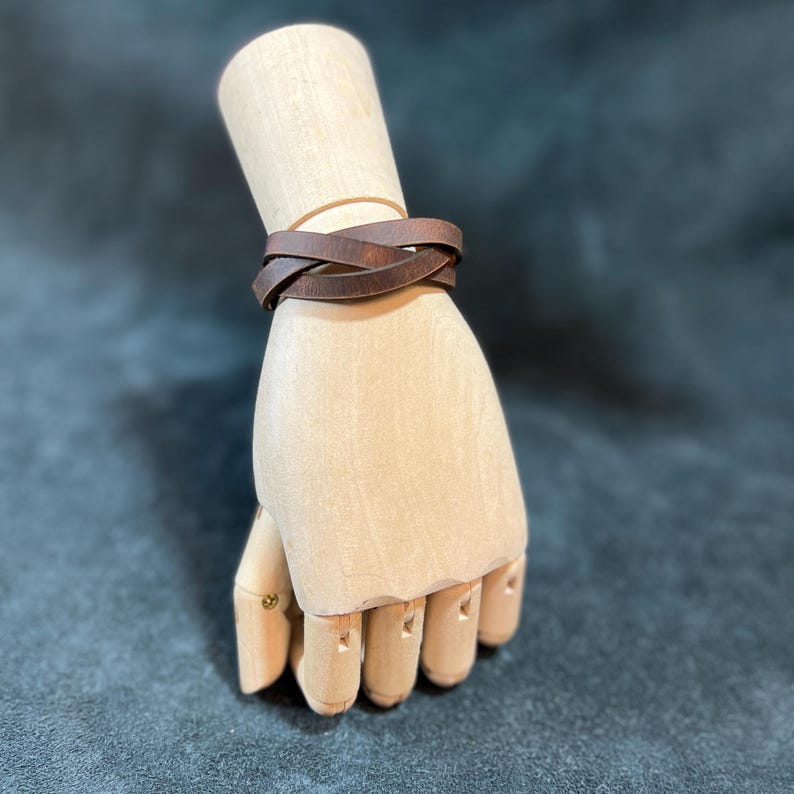 May include: A dark brown leather bracelet featuring a simple wrap-around design with a crossed detail. The bracelet is displayed on a light-colored wooden mannequin hand, showcasing the smooth texture and matte finish of the leather material.