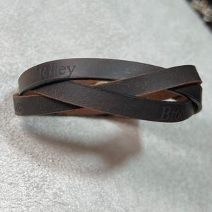 May include: A dark brown leather wrap bracelet featuring a crisscross design. The leather band is embossed with the names Riley and Brook in a serif font. The material has a matte, textured finish with visible natural grain throughout the surface.