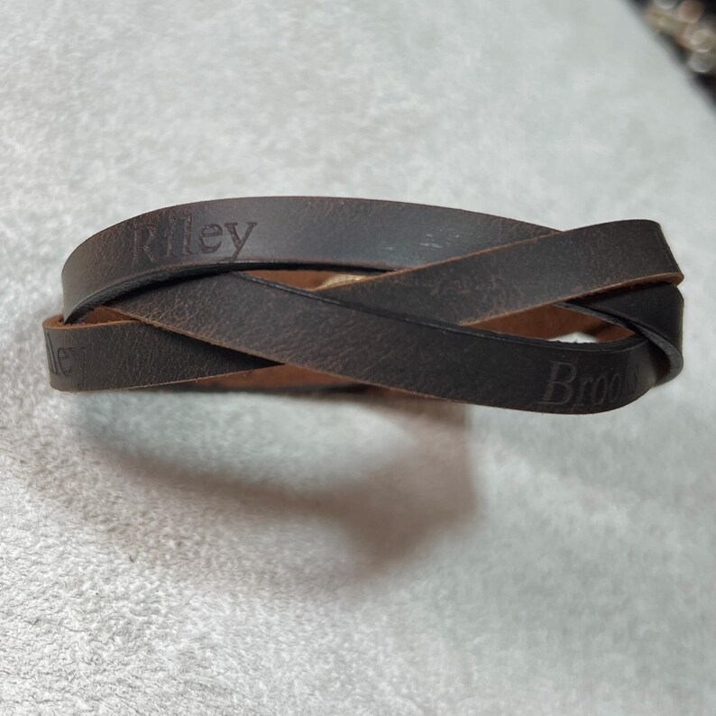 May include: A dark brown leather wrap bracelet featuring a crisscross design. The leather band is embossed with the names Riley and Brook in a serif font. The material has a matte, textured finish with visible natural grain throughout the surface.