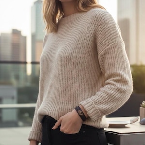 Puede incluir: Un su&eacute;ter de punto beige con detalles acanalados en los pu&ntilde;os y el dobladillo. El su&eacute;ter tiene un corte holgado y mangas largas. Una pulsera de cuero negro con un dise&ntilde;o dorado es visible en la mu&ntilde;eca. La persona lleva pantalones negros.