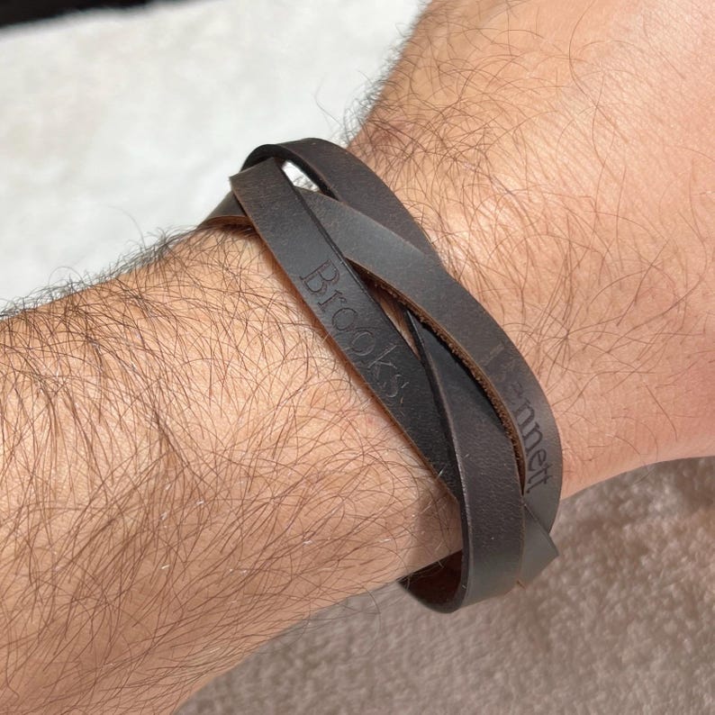 May include: A dark brown leather wrap bracelet featuring two overlapping bands. The leather surface is engraved with the names Brooks and Bennett in a serif font. The bracelet is designed to wrap around the wrist, creating a layered appearance.