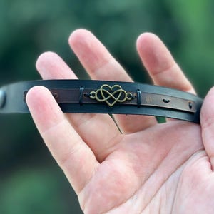 Puede incluir: Pulsera de cuero negro con un acento de cuero marr&oacute;n y un dije de coraz&oacute;n infinito de color bronce. La pulsera se sostiene en una mano, mostrando los detalles del dise&ntilde;o. Un accesorio elegante.