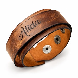 May include: A brown leather wrap bracelet featuring a distressed finish and the name Alicia engraved in a black script font. The bracelet includes two metallic snap buttons on the interior for closure, securing the layered leather band design.
