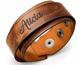 May include: A brown leather wrap bracelet featuring a distressed finish and the name Alicia engraved in a black script font. The bracelet includes two metallic snap buttons on the interior for closure, securing the layered leather band design.