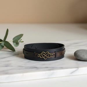 Puede incluir: Pulsera de cuero negra con un acento de cuero marr&oacute;n y un s&iacute;mbolo de infinito en forma de coraz&oacute;n de color dorado. La pulsera se muestra sobre una superficie de m&aacute;rmol blanco.