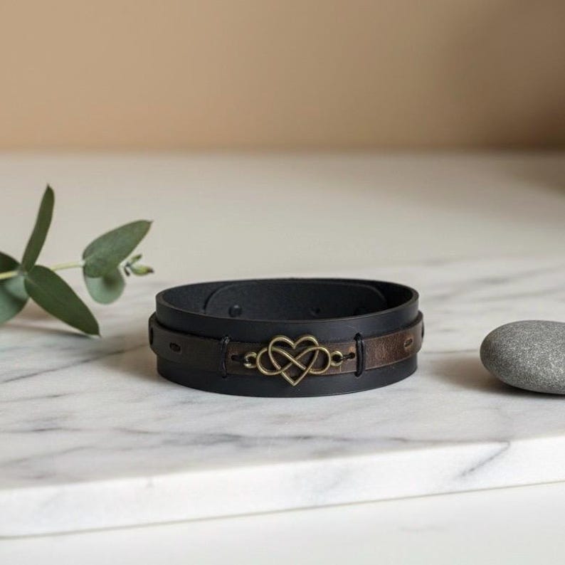 Puede incluir: Pulsera de cuero negra con un acento de cuero marr&oacute;n y un s&iacute;mbolo de infinito en forma de coraz&oacute;n de color dorado. La pulsera se muestra sobre una superficie de m&aacute;rmol blanco.