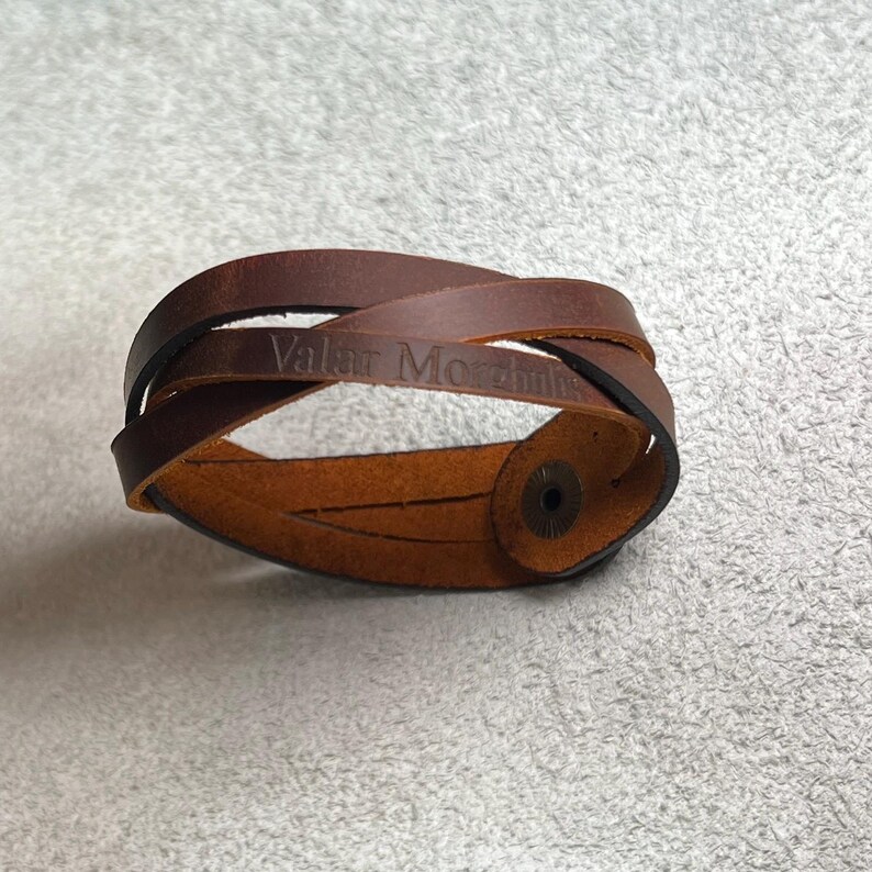 May include: A brown leather wrap bracelet featuring a crisscross design and a metal snap button closure. The surface of the leather is embossed with the text Valar Morghulis in a dark font, creating a clear contrast against the brown material.