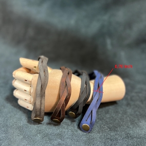 May include: Four braided leather bracelets in gray, brown, black, and blue. Each bracelet features a metallic snap button closure and a width of 0.70 inches as indicated by the red text and line pointing to the blue bracelet's strap.