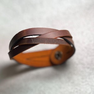 May include: A brown leather bracelet featuring a braided design with the text Valar Morghulis embossed on the surface. The bracelet is secured with a metallic snap button closure, showcasing a smooth finish and a natural tan interior lining.