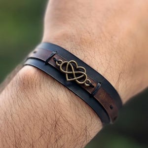 Puede incluir: Una pulsera de cuero con un s&iacute;mbolo de infinito en forma de coraz&oacute;n de bronce. La pulsera tiene una banda negra y una banda marr&oacute;n, cosidas juntas. La pulsera est&aacute; dise&ntilde;ada para usarse en la mu&ntilde;eca.