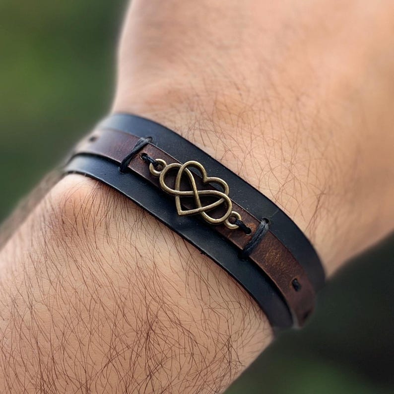 Puede incluir: Una pulsera de cuero con un s&iacute;mbolo de infinito en forma de coraz&oacute;n de bronce. La pulsera tiene una banda negra y una banda marr&oacute;n, cosidas juntas. La pulsera est&aacute; dise&ntilde;ada para usarse en la mu&ntilde;eca.
