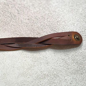 May include: A brown leather bracelet featuring a braided design and a metallic snap button closure. The leather surface is embossed with the text Valar Morghulis, showing a smooth finish with natural variations in the brown color tone.