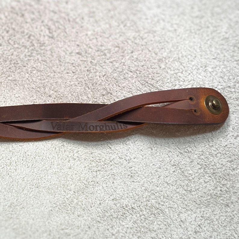 May include: A brown leather bracelet featuring a braided design and a metallic snap button closure. The leather surface is embossed with the text Valar Morghulis, showing a smooth finish with natural variations in the brown color tone.