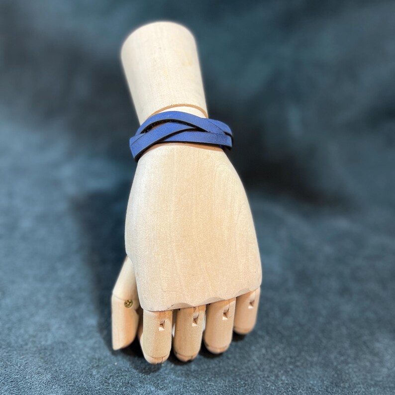 May include: A dark blue bracelet featuring a braided design made of multiple thin, flat strips. The bracelet is wrapped around a light-colored wooden mannequin hand, showcasing the smooth texture and the layered, intertwined structure of the band.