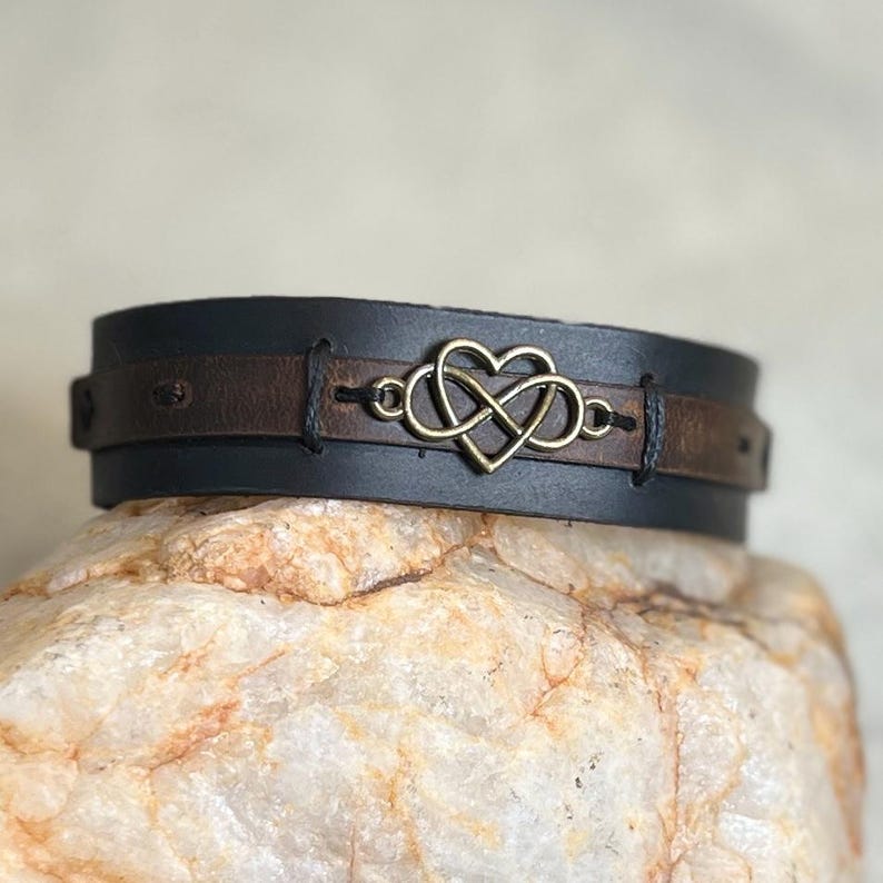 Puede incluir: Una pulsera de cuero negro y marr&oacute;n con un s&iacute;mbolo de infinito en forma de coraz&oacute;n de bronce. La pulsera est&aacute; hecha de dos capas de cuero, con el s&iacute;mbolo de bronce en el centro. La pulsera es de estilo brazalete.
