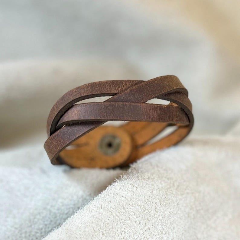 May include: A brown leather bracelet featuring multiple overlapping straps that create a layered, crisscross design. The bracelet is secured with a circular metal snap fastener visible on the inner side of the band, showcasing a smooth, worn texture.