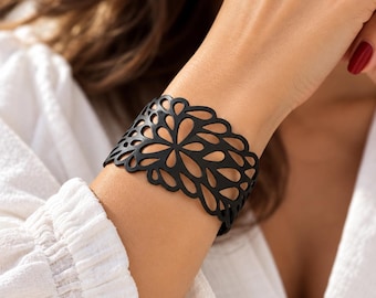 Puede incluir: Una pulsera de brazalete ancha de color negro que presenta un intrincado patrón floral cortado con láser con formas repetitivas de pétalos. La pulsera tiene un acabado mate suave y envuelve la muñeca, mostrando un delicado diseño calado en toda la banda.