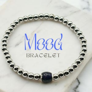 May include: A silver beaded bracelet with a dark blue central bead. The word "Mood" is written in blue script above the word "BRACELET" in a sans-serif font. The bracelet is displayed on a white marble surface.