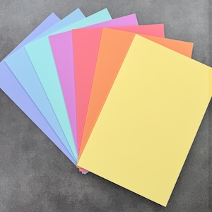 May include: A selection of colourful cardstock paper in shades of lavender, light blue, teal, pink, orange, and yellow. The sheets are arranged in a layered, fanned-out pattern, showcasing the variety of colours. The paper is smooth and appears to be of a standard size.