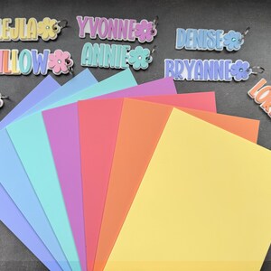 May include: A collection of colorful name tags with flower accents and a variety of pastel-colored paper sheets. The name tags include names like Lejla, Yvonne, Denise, Willow, Annie, Bryanne, Jules, and Lorena.