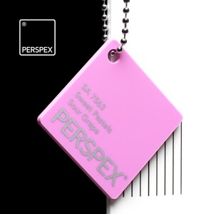 May include: A square, pink Perspex color swatch, suspended by a silver ball chain. The swatch features the text "SA 7563 Sweet Pastels Sour Grape" and the brand name "PERSPEX" in silver. The background is split, with black on the left and white on the right.