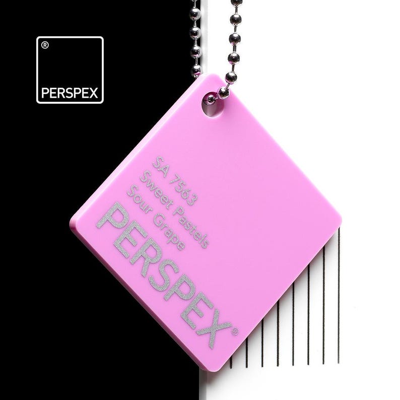 May include: A square, pink Perspex color swatch, suspended by a silver ball chain. The swatch features the text "SA 7563 Sweet Pastels Sour Grape" and the brand name "PERSPEX" in silver. The background is split, with black on the left and white on the right.