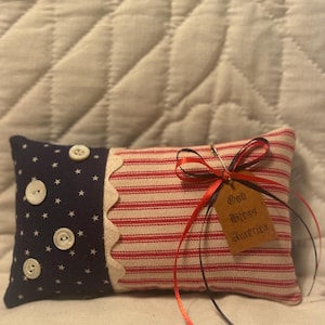 Op de afbeelding: Een rechthoekig kussen met een marineblauw gedeelte met sterrenpatroon en een rood-wit gestreept gedeelte. Het heeft witte knopen, een geschulpte rand en een label met de tekst "God Bless America."