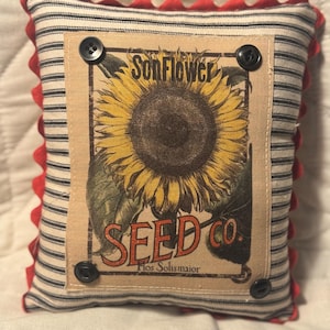 May include: A decorative pillow with a vintage sunflower seed packet design. The pillow features a striped fabric with red trim and four black buttons. The sunflower image includes the text "SonFlower SEED CO."