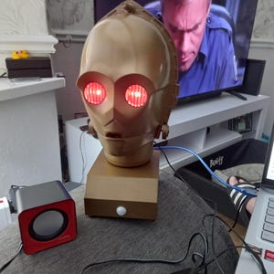 May include: A gold-coloured C-3PO head with glowing red eyes sits on a wooden base. Two small black and red speakers are connected. A television screen is visible in the background.