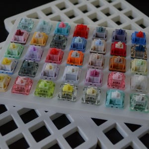 May include: A collection of colorful mechanical keyboard switches arranged on a white grid. The switches are various colors, including red, blue, yellow, and green. The image is well-lit, showcasing the details of each switch.
