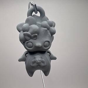 Puede incluir: Un llavero gris impreso en 3D con un personaje de dibujos animados estilizado con ojos grandes y una superficie texturizada. El personaje tiene un cuerpo redondo y un lazo para sujetar. Ideal para personalizar llaves o bolsos.