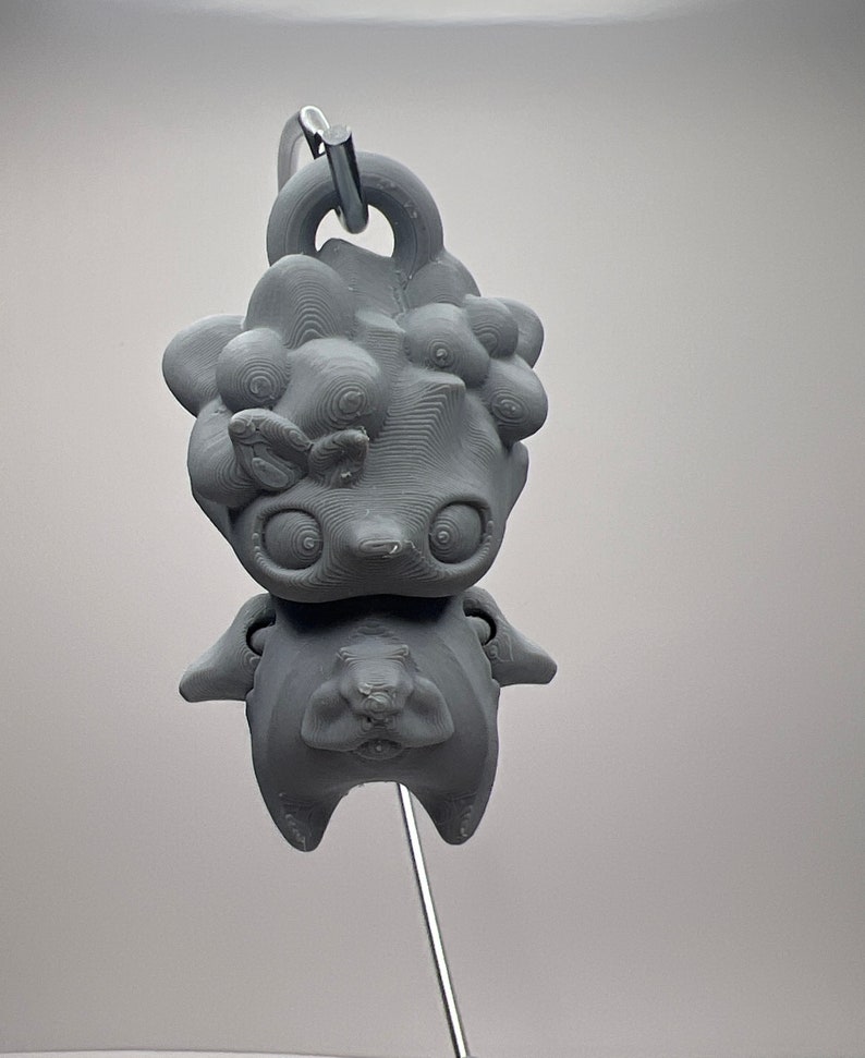 Puede incluir: Un llavero gris impreso en 3D con un personaje de dibujos animados estilizado con ojos grandes y una superficie texturizada. El personaje tiene un cuerpo redondo y un lazo para sujetar. Ideal para personalizar llaves o bolsos.