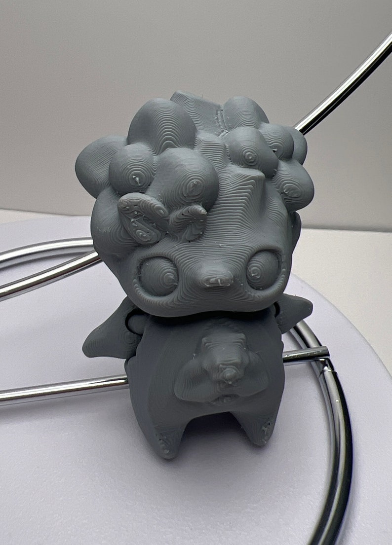 Puede incluir: Una figura gris impresa en 3D de una criatura de dibujos animados con ojos grandes y una superficie texturizada. La figura tiene una cabeza redondeada con rasgos similares a burbujas y brazos peque&ntilde;os. Se encuentra sobre una superficie blanca.