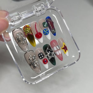 Puede incluir: Una caja de plástico transparente con 12 uñas postizas de colores. Las uñas están decoradas con varios diseños, incluyendo estrellas, dados, cerezas y cadenas.