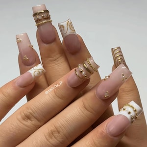 May include: A hand with long, square-shaped acrylic nails. The nails are painted in a nude color with gold accents, including small beads and rings. Some nails feature white tips with gold swirl designs. The text "@nails.by_jk" is visible.