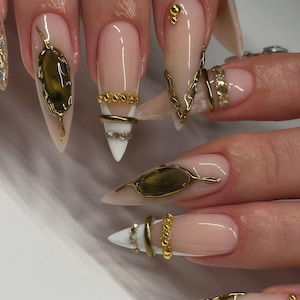 May include: Close-up of long, pointed acrylic nails with a nude base and gold embellishments. Designs include gold leaf shapes, chain details, and ring accents with rhinestones. The nails have a glossy finish.