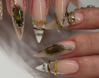 Puede incluir: Primer plano de uñas acrílicas largas y puntiagudas con base nude y adornos dorados. Los diseños incluyen formas de hojas doradas, detalles de cadenas y acentos de anillos con pedrería. Las uñas tienen un acabado brillante.