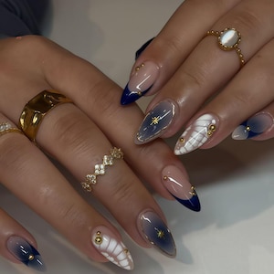 May include: Close-up of manicured nails with diverse designs. Some nails have a blue and grey gradient, others feature white and gold accents. Gold rings are visible on the fingers.