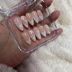 May include: A set of ten almond-shaped press-on nails with a nude base color and intricate white floral and leaf line art designs. The nails are arranged in a clear plastic storage case with a hinged lid, showcasing various botanical patterns.