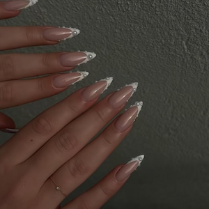 May include: Close-up of manicured nails with a nude base and white tips. The tips are adorned with small, white pearls. A delicate gold ring with a small diamond is visible on one finger. The nails are long and almond-shaped.