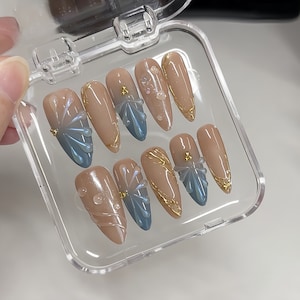 May include: A set of ten nude-colored press-on nails with blue and gold accents. The nails have a floral design with gold glitter and blue swirls.