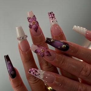 Puede incluir: Primer plano de una mano con uñas largas pintadas de rosa y blanco con diseños florales y abstractos. Las uñas están decoradas con pedrería y detalles dorados.