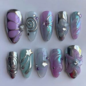 May include: A collection of ten artificial nails with diverse designs. The nails showcase silver, purple, and iridescent colours. Some nails feature butterfly, swirl, and star designs, while others have floral embellishments.