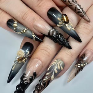 Puede incluir: Primer plano de un conjunto de uñas acrílicas largas y puntiagudas. Las uñas presentan una variedad de diseños, que incluyen efectos ombre, adornos 3D y patrones intrincados. Los colores incluyen negro, marrón, beige y dorado, con pedrería y detalles florales.