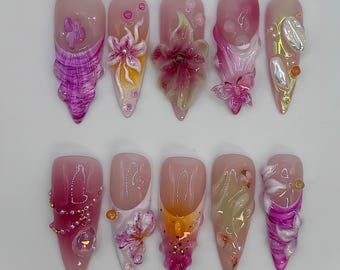 May include: Ten pink and white stiletto shaped fake nails with various 3D floral designs, including butterflies, flowers, and swirls.
