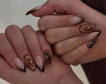May include: Almond-shaped nails with a gradient effect, transitioning from a nude base to a deep brown at the tips. Gold accents include a thin outline and starburst designs, creating a sophisticated and elegant manicure.