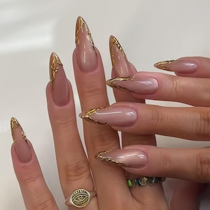 May include: Long, almond-shaped nails with a nude base and gold metallic tips. The nails are adorned with a delicate gold design. A silver ring with an eye symbol is visible on one finger.